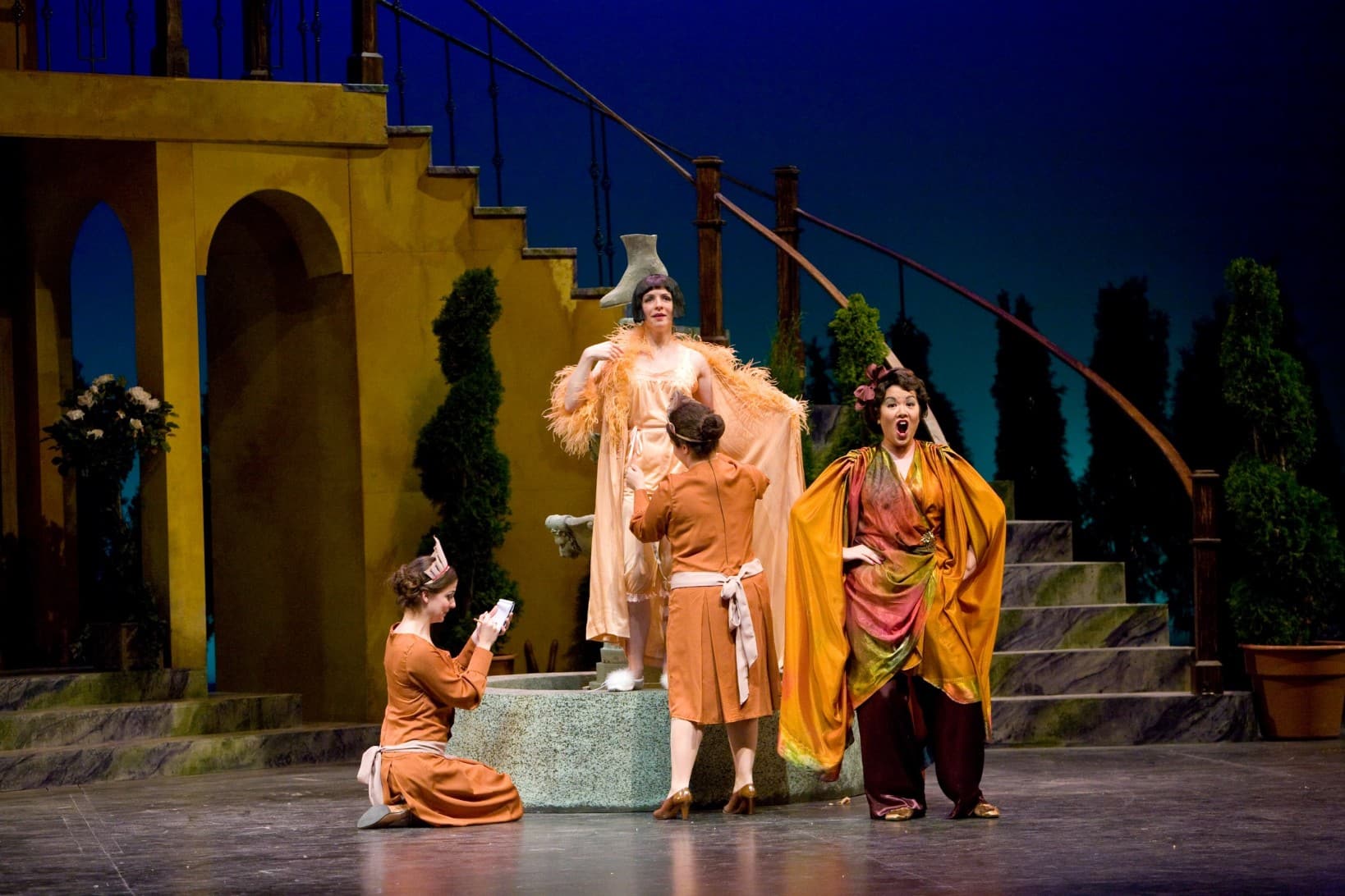 This image contains the same characters as the other photos. While the characters in the other photos were consistently dressed
                            in each picture, they are wearing more decorative clothes in this scene. Sarah is standing on the fountain with a feathered
                            boa, while the woman who wear red is standing in front of her in a yellow cape and singing.
