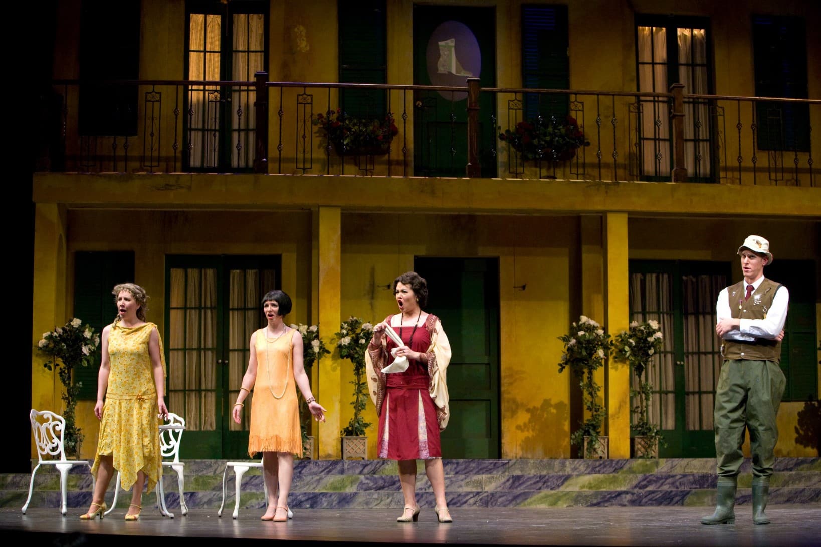 Three women singing in unison on stage, two in yellow dresses and one wearing red. A man on stage in a vest watches on the
                            side.