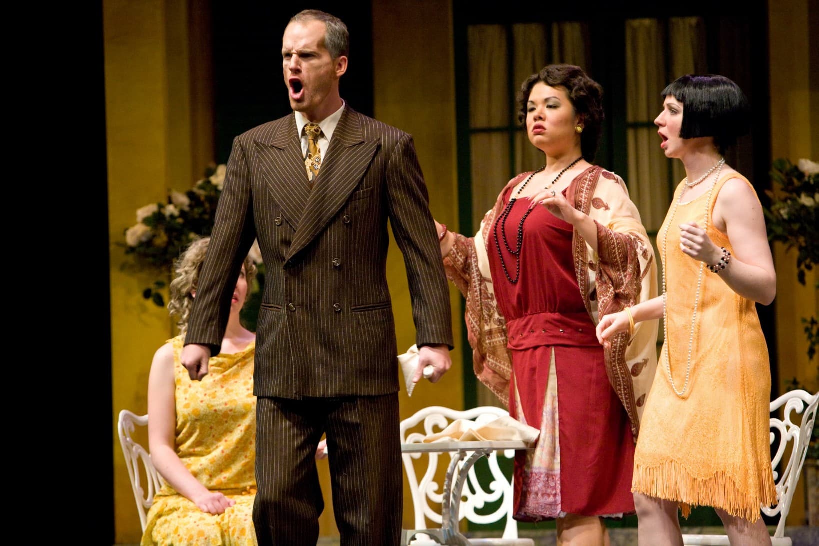 A man is singing with an assertive look on his face. An equally-assertive woman stands behind him, while a third woman is
                            on the side looking concerned.