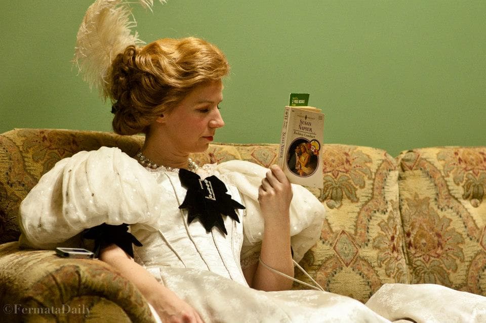 A woman is wearing a turn of the century dress with large poofy shoulder pads, a corset, and a plume in her hair. She is
                            reading the back of a book.