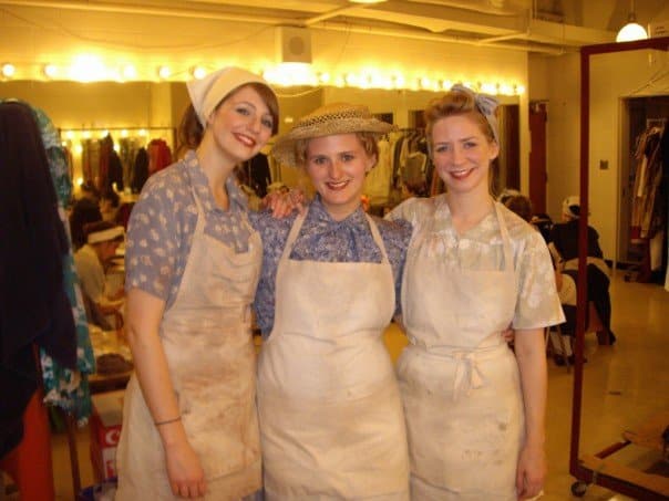 Three women are standing in the middle of a dressing room in costume - they are wearing chef's aprons.