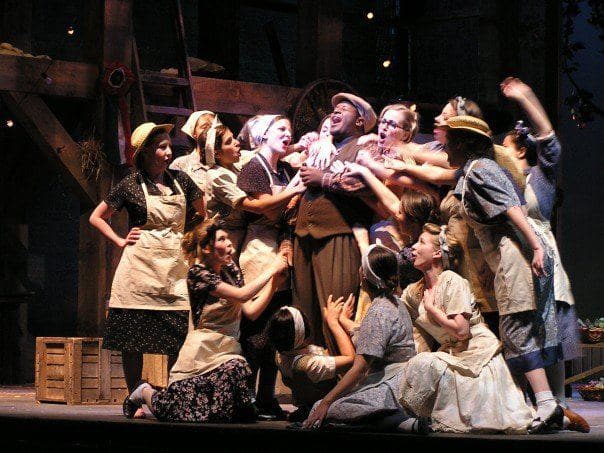 A man is center stage, singing to the sky, and around him there are around 10 women singing with him, propping him up, and
                            embracing him in an uplifting manner. The light is shining directly in his face, giving the scene a look of hope for the
                            future.