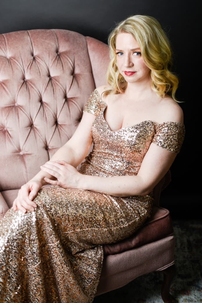 Sarah is in her gold sequins dress and sitting on the pink velvet couch. The dress has sleeves that go off the shoulder,
                            and the length goes down to her feet.
