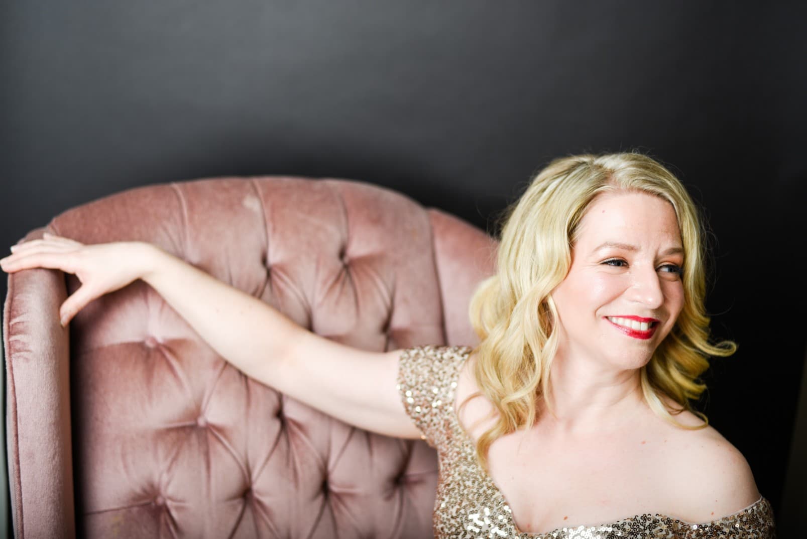 Sarah is in the gold sequins dress and sitting on the couch. she has her right arm on the couch, while her body is centered
                            and her face is looking to her left.