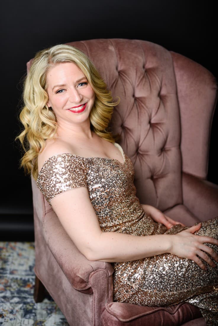 Sarah is sitting on the pink velvet couch in her gold sequins dress. There is a faded colourful carpet on the floor, and
                            she is looking at the camera with her body facing away to the right of the frame.