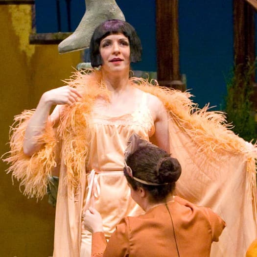 This image contains the same characters as the other photos. While the characters in the other photos were consistently dressed in each picture, they are wearing more decorative clothes in this scene. Sarah is standing on the fountain with a feathered boa, while the woman who wear red is standing in front of her in a yellow cape and singing.