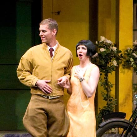 Sarah at center stage in a black wig next to a man. Two women are standing on either side of them, one with a look of amusement on her face.