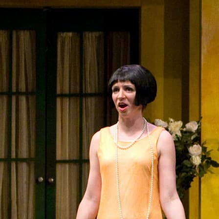 Three women singing in unison on stage, two in yellow dresses and one wearing red. A man on stage in a vest watches on the side.