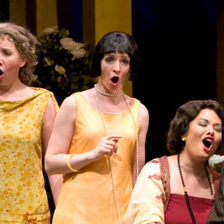6 people on stage, singing in unison. Two are seated on a fountain while the rest are standing around them.