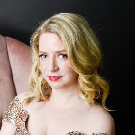 Sarah is in her gold sequins dress and sitting on the pink velvet couch. The dress has sleeves that go off the shoulder, and the length goes down to her feet.