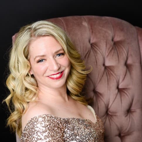 Sarah is sitting on the pink velvet couch in her gold sequins dress. There is a faded colourful carpet on the floor, and she is looking at the camera with her body facing away to the right of the frame.