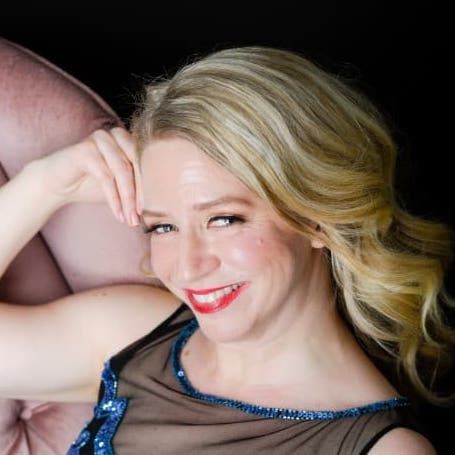 Sarah is wearing her blue sequins dress, leaning back on a pink velvet couch, and smiling up at the camera.