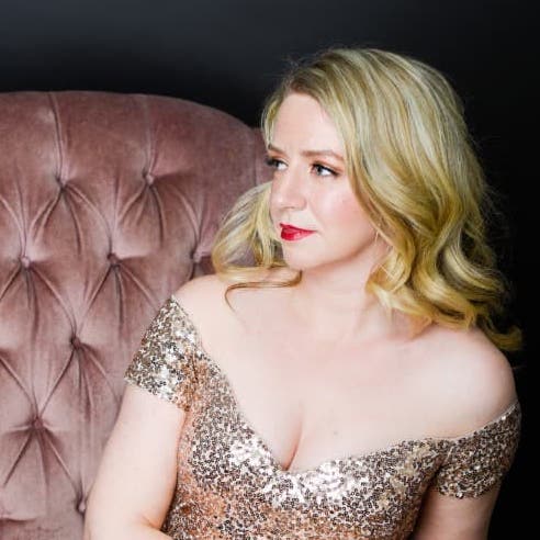 Sarah is sitting in her gold sequins dress. Her legs are crossed and she is looking to her right in a neutral manner.