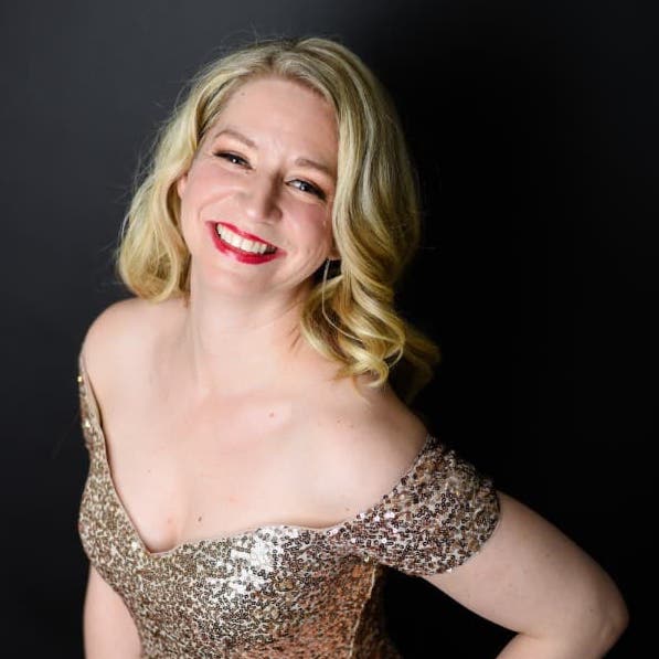 Sarah is wearing her gold sequins dress. The camera is higher than her. Her left hand is on her waist and she is smiling up at the camera.