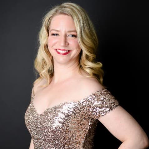 Sarah is standing in her gold sequins dress. He background is black and her fair skin, hair, and dress all shine against it. She is smiling at the camera with a hand on her waist.
