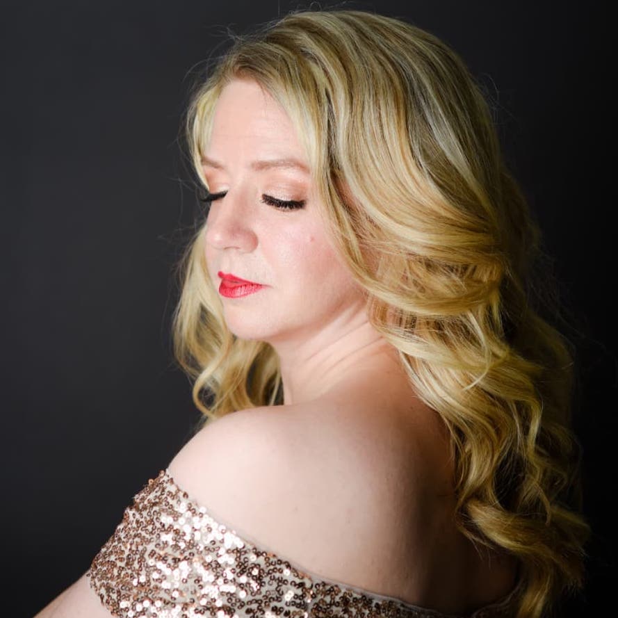 Sarah is wearing a gold sequins dress. The image just shows above her shoulders, and she is turned away from the camera. Her face is turned slightly back, and she is looking down with soft eyes.