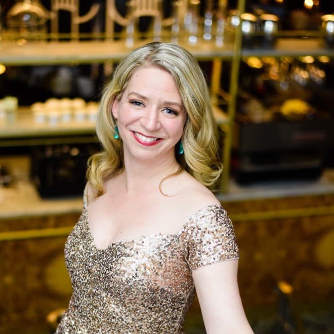 Sarah is in her gold sequins dress, holding on to a railing behind her. She is standing on the balcony of a performace hall, and you can see the other balconies out of focus behind her.