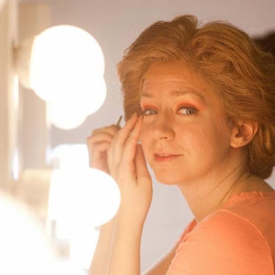 A woman is sitting in front of a well-lit mirror doing he makeup. The photo is snapped as she looks over her shoulder at the camera, mid makeup application.