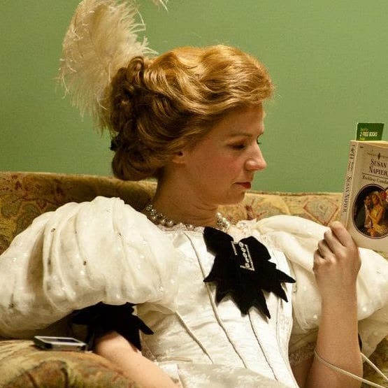 A woman is wearing a turn of the century dress with large poofy shoulder pads, a corset, and a plume in her hair. She is reading the back of a book.