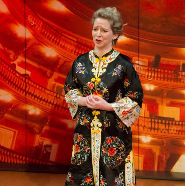 Sarah is standing on stage, wearing a frock in a traditional Chinese style with gold sleeves and lots of embroidery.