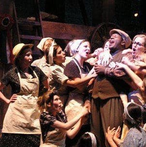 A man is center stage, singing to the sky, and around him there are around 10 women singing with him, propping him up, and embracing him in an uplifting manner. The light is shining directly in his face, giving the scene a look of hope for the future.